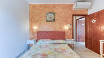 A bedroom with a tufted headboard, a bed with a colorful circular-patterned bedspread, two bedside tables, a wall-mounted air conditioner, and a door with a diamond-shaped decoration.