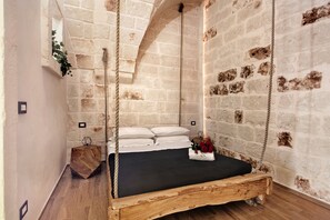 Une pièce rustique avec un lit-hamac en bois, des murs en briques apparentes et une petite table d’appoint en bois.