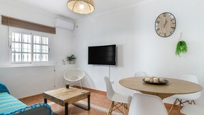 A modern living room with a flat-screen TV, a round wooden table, white chairs, a sofa, and a large wall clock.