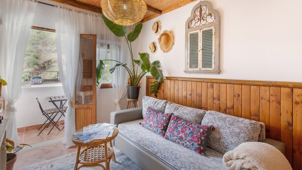 Una habitación acogedora con techo de madera, sofá con cojines estampados, lámpara colgante de mimbre y ventana con contraventanas.