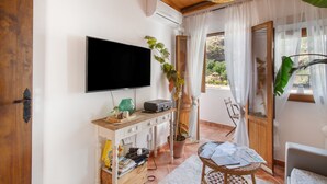 Una habitación con una televisión de pantalla plana montada en la pared, un mueble de madera con varios objetos, una planta en maceta, una silla de mimbre y una mesa con papeles.