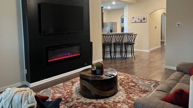 Un salon avec une cheminée, un ventilateur de plafond et une cuisine en arrière-plan.