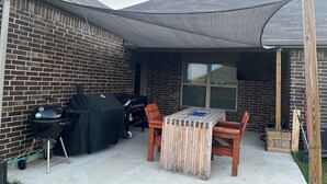 Une terrasse à l’arrière de la maison, avec un barbecue, une table couverte et une chaise en bois.