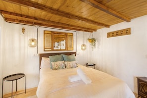 A bedroom with a wooden ceiling, a bed with floral pillows, a bedside table, and a hanging plant.