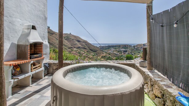 A hot tub with a view of a valley and mountains, a barbecue area, and a pergola with string lights.