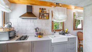 Een keuken met een fornuis, magnetron en spoelbak. Er zijn kastjes, een raam en een stoel.