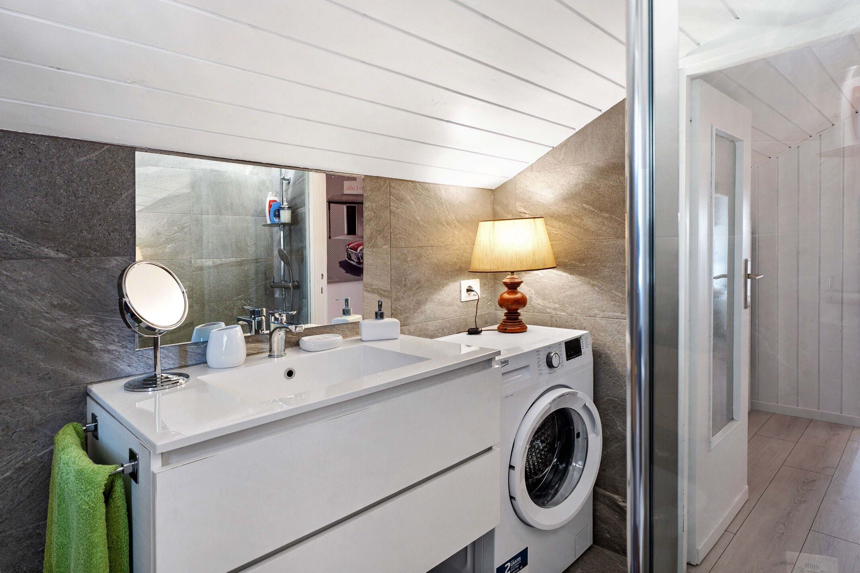 A modern bathroom with a large mirror, dual sinks, a washing machine, and a lamp.