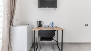 Ein kleines Zimmer mit Schreibtisch, Stuhl, Fernseher, Kühlschrank und Wasserkocher.