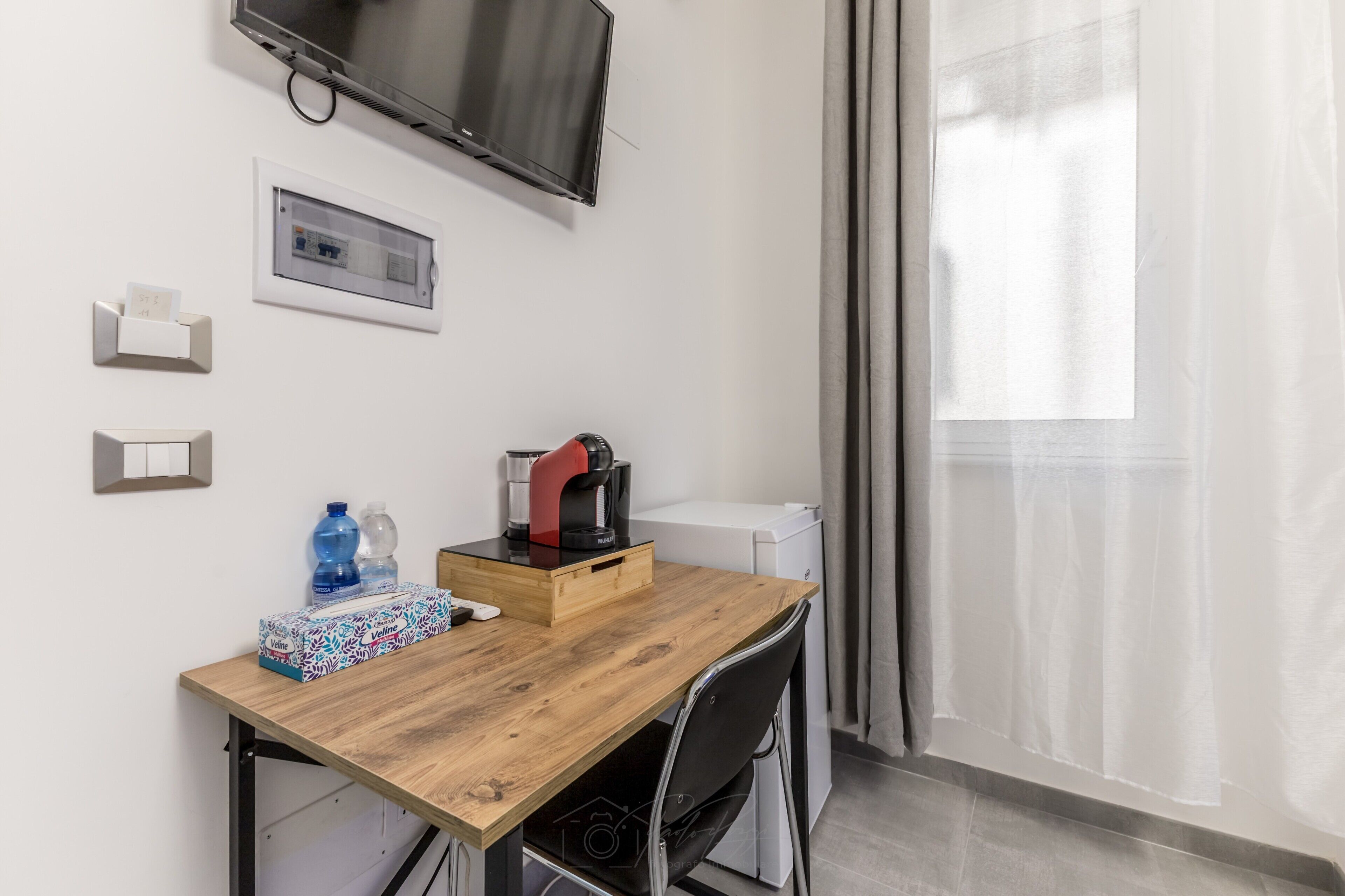 A small room with a wooden table, a TV mounted on the wall, a coffee maker, a water dispenser, and a window with curtains.