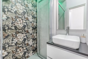 Un baño con papel tapiz floral, cabina de ducha de vidrio, lavabo blanco y un espejo.