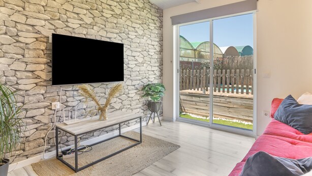 A living room with a stone wall, a flat-screen TV, a wooden bench, a red sofa, and a sliding glass door leading to a patio. - Cottage 'Casita de Ana' with Private Pool, Wi-Fi and Air Conditioning (Conil de la Frontera)