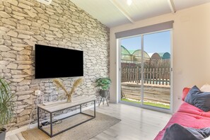 A living room with a stone wall, a flat-screen TV, a wooden bench, a red sofa, and a sliding glass door leading to a patio.