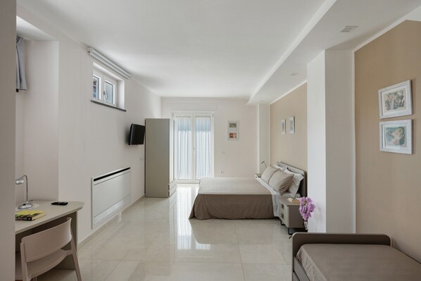 A hotel room with a bed, a desk with a chair, a TV, a refrigerator, and a window with blinds.