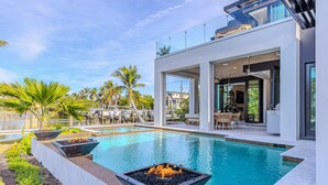 A modern outdoor pool area with a fire feature, lounge chairs, and a glass-enclosed patio. - NEW LUX WATERFRONT HOME- Walk to 5th Ave (Naples)