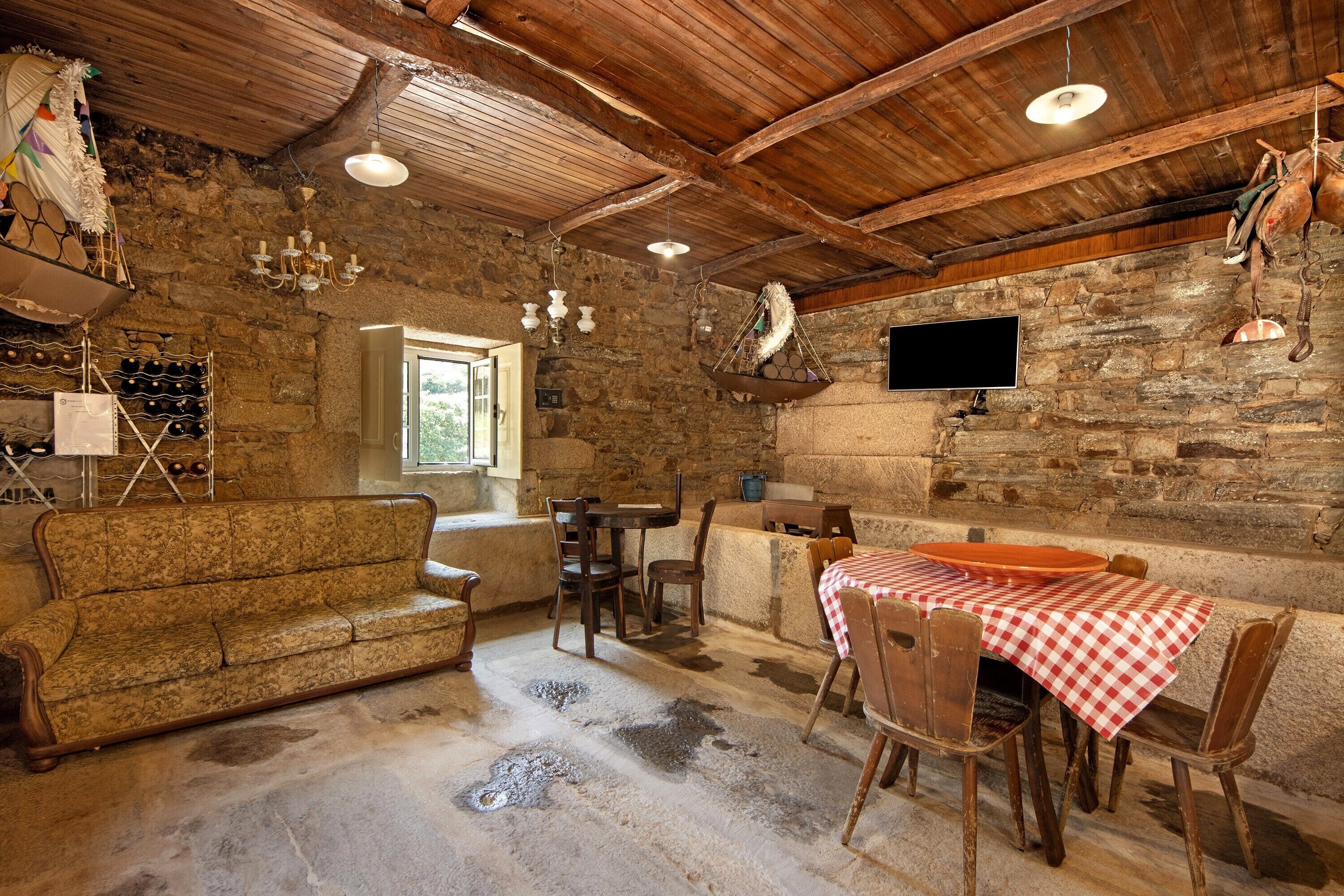 Ein rustikales Interieur mit Steinwand, Holzbalkendecke, einer Couch, einem Tisch mit kariertem Tischlein und einem an der Wand befestigten Fernseher.