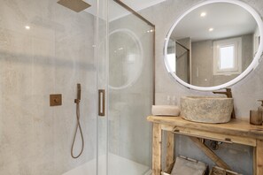 A modern bathroom with a glass shower enclosure, a wooden vanity with a stone basin, and a round mirror.
