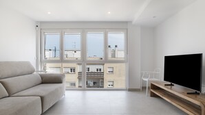 Una sala de estar moderna con un sofá, un televisor sobre un soporte de madera y ventanas grandes con vistas a edificios.