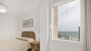 Un dormitorio con cama, mesita de noche de madera, ventana con vista al mar y edificios, y dos cuadros en la pared.
