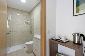 Une salle de bain moderne avec une douche à parois de verre, des toilettes blanches, un meuble-lavabo en bois et une bouilloire posée sur une petite table d’appoint en bois.