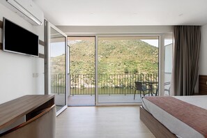 A hotel room with a bed, a TV, a balcony with a table and chairs, and a view of a hilly landscape.