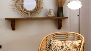 Une chaise en osier avec un coussin à motifs floraux, un miroir fixé au mur, une lampe sur pied et une étagère ornée d’objets décoratifs.