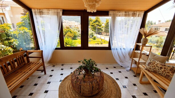 Une pièce avec un banc en bois, une table basse en osier et une vue sur la verdure et les bâtiments, grâce à de grandes fenêtres ornées de rideaux légers.