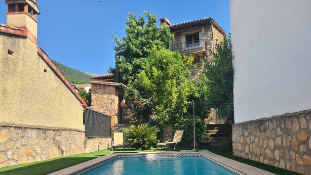 Una piscina con un muro de piedra, un área verde y un edificio con balcón.