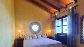Un dormitorio con techo de madera, una cama con cubrecama blanco, dos lámparas de noche, un adorno circular en la pared, una ventana con cortinas y un cofre de madera.