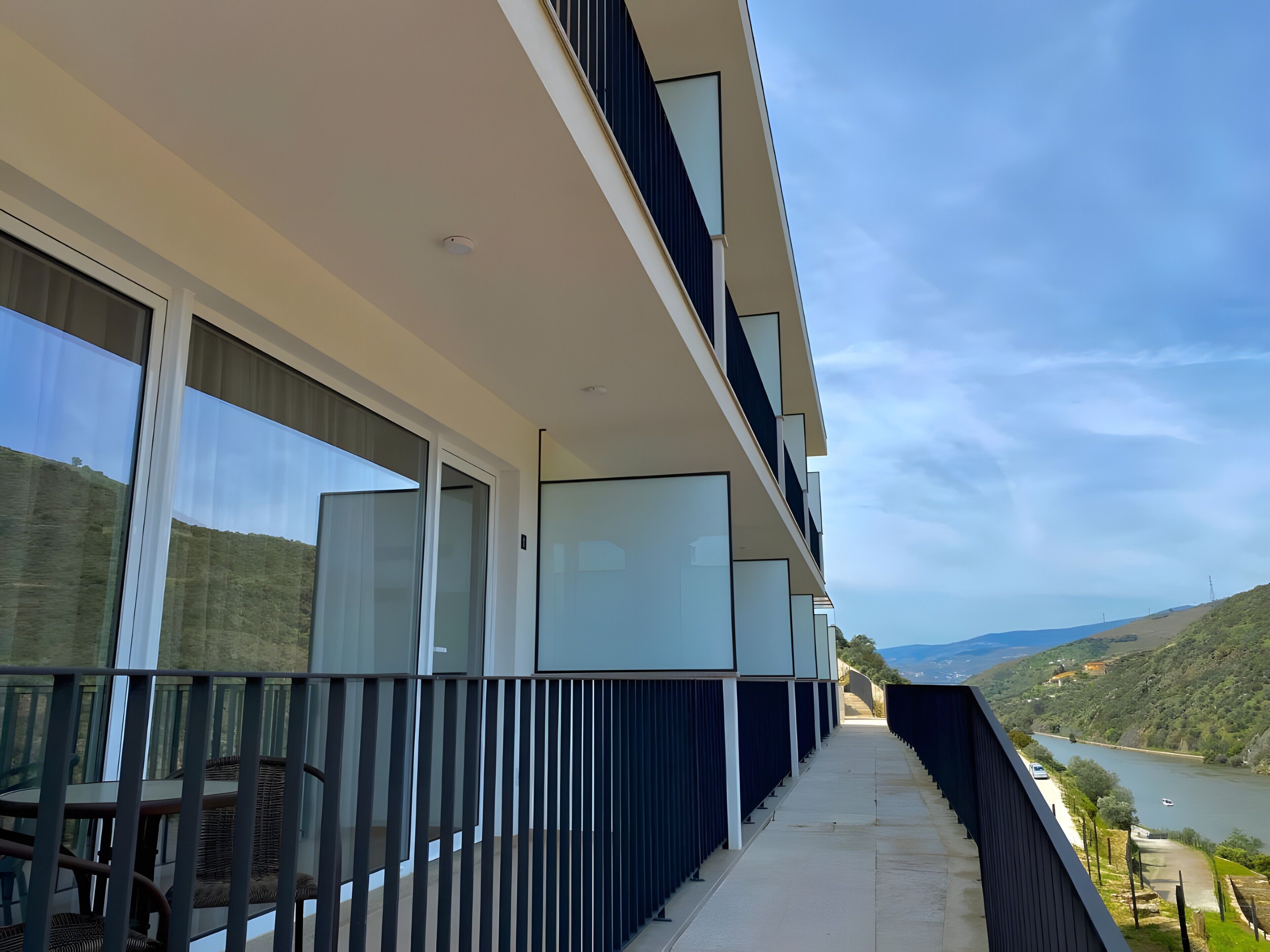 Un bâtiment moderne doté de grandes fenêtres, d’un balcon agrémenté de meubles en osier et offrant une vue sur une rivière et des collines.