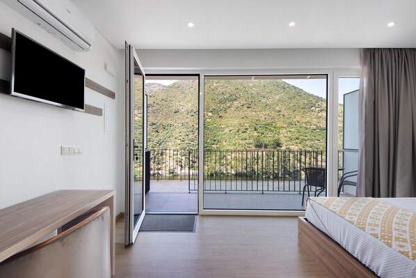 Habitación de hotel con cama, escritorio, televisión y balcón con vistas a un paisaje montañoso.