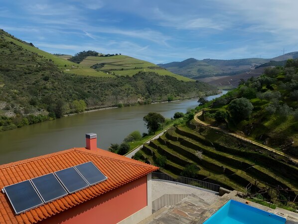 Un viñedo en terrazas con un río, un edificio de techo rojo con paneles solares y una piscina.