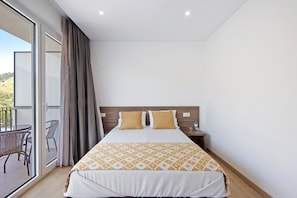Ein Hotelzimmer mit einem Bett, einem Nachttisch, einer Glastür, die auf einen Balkon führt, und einem Sessel.