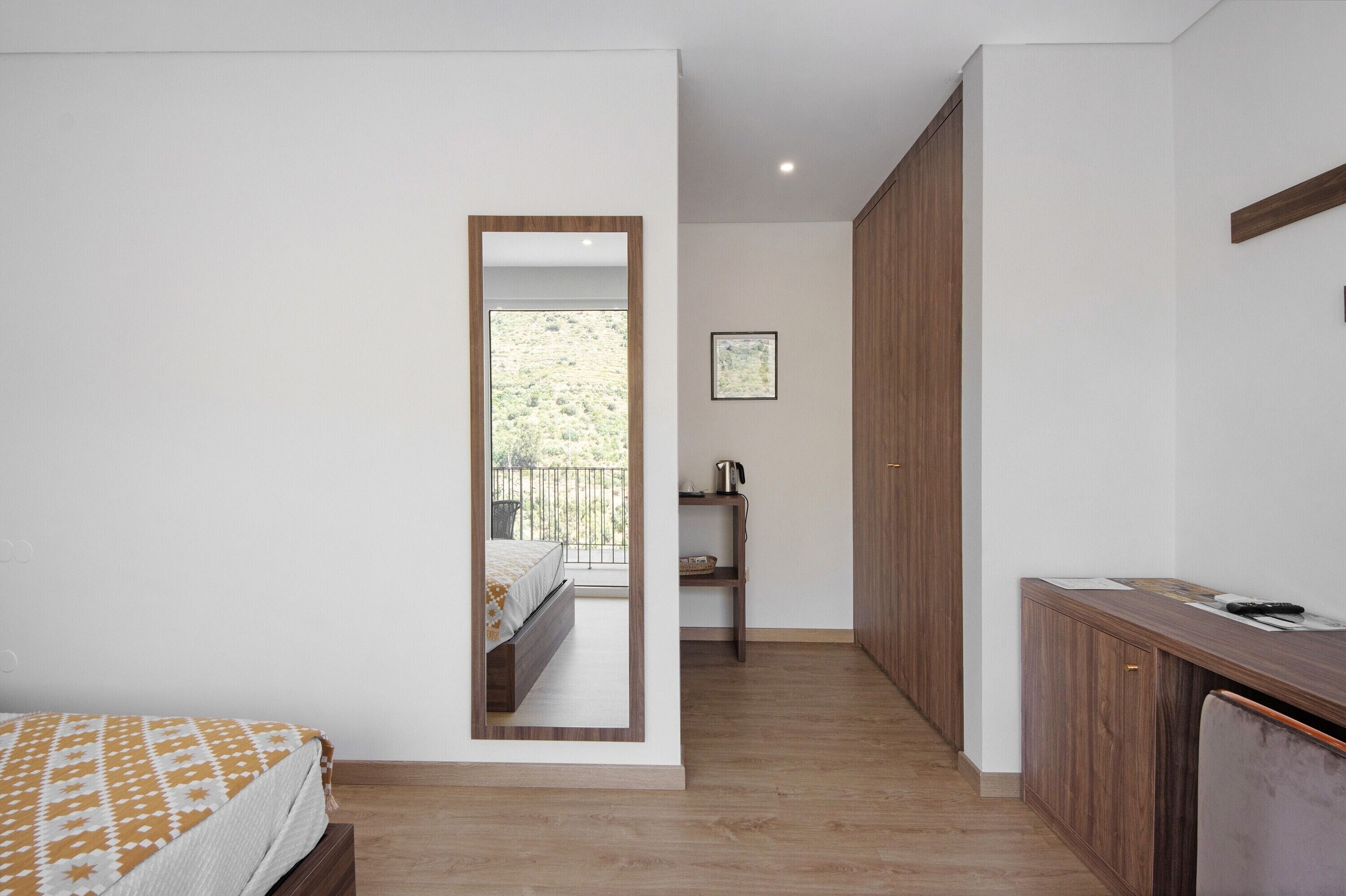 A bedroom with a bed, a wooden wardrobe, a desk with a chair, a small shelf with a kettle, and a framed picture on the wall.