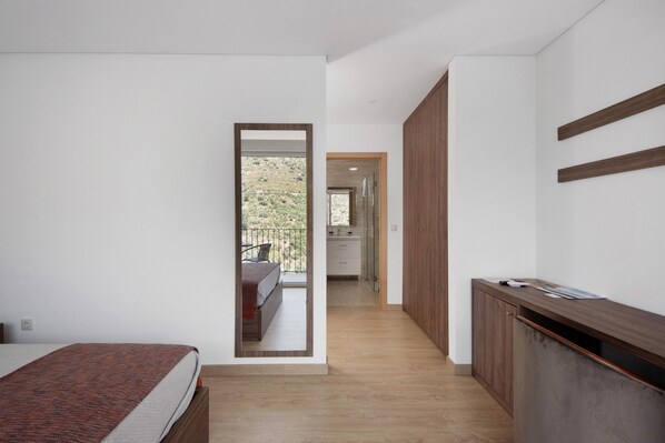 A bedroom with a bed, a wooden desk, a mirror, and a view of a balcony.