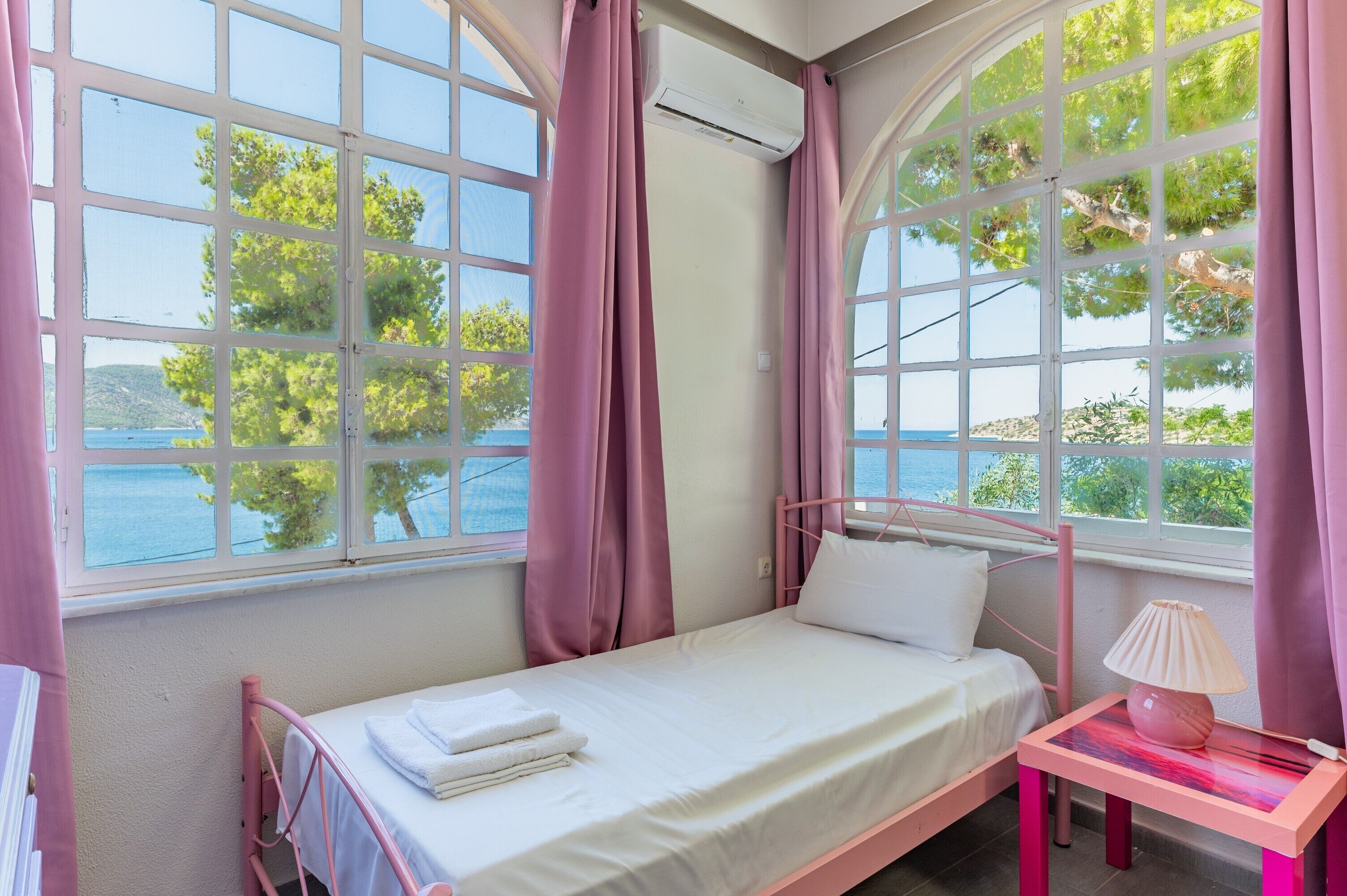 Una cama individual con colchón y almohadas blancas, una mesita de noche con lámpara, y ventanas grandes con cortinas rosadas.