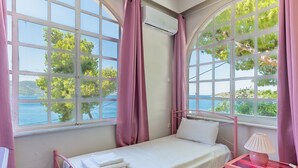 A single bed with a white mattress and pillows, a bedside table with a lamp, and large windows with pink curtains. - Apartment 'Afroditi' with Sea View, Wi-Fi and Air Conditioning (Salamina)