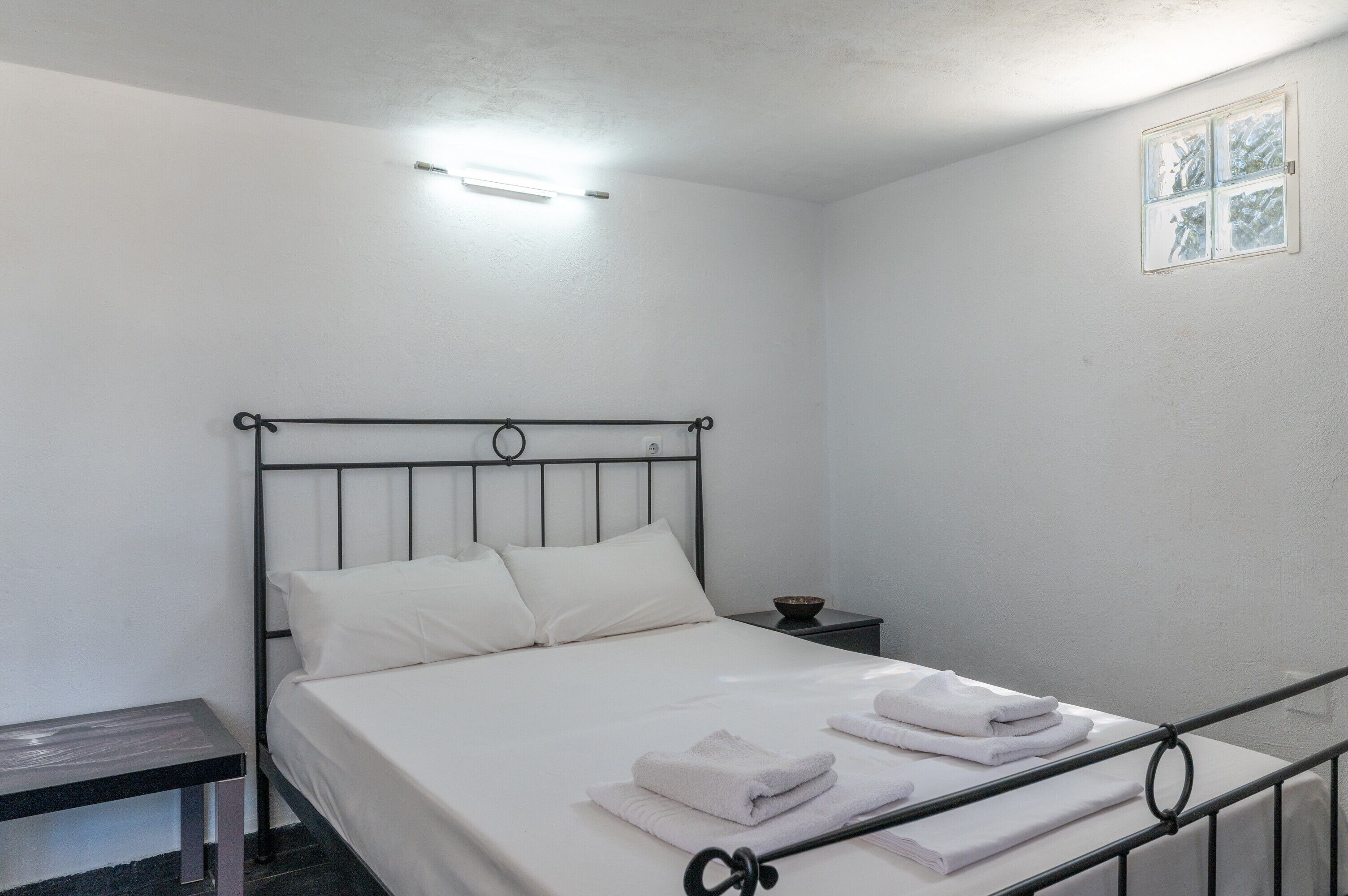 A single bed with white bedding, a bedside table, and a small window with blinds.