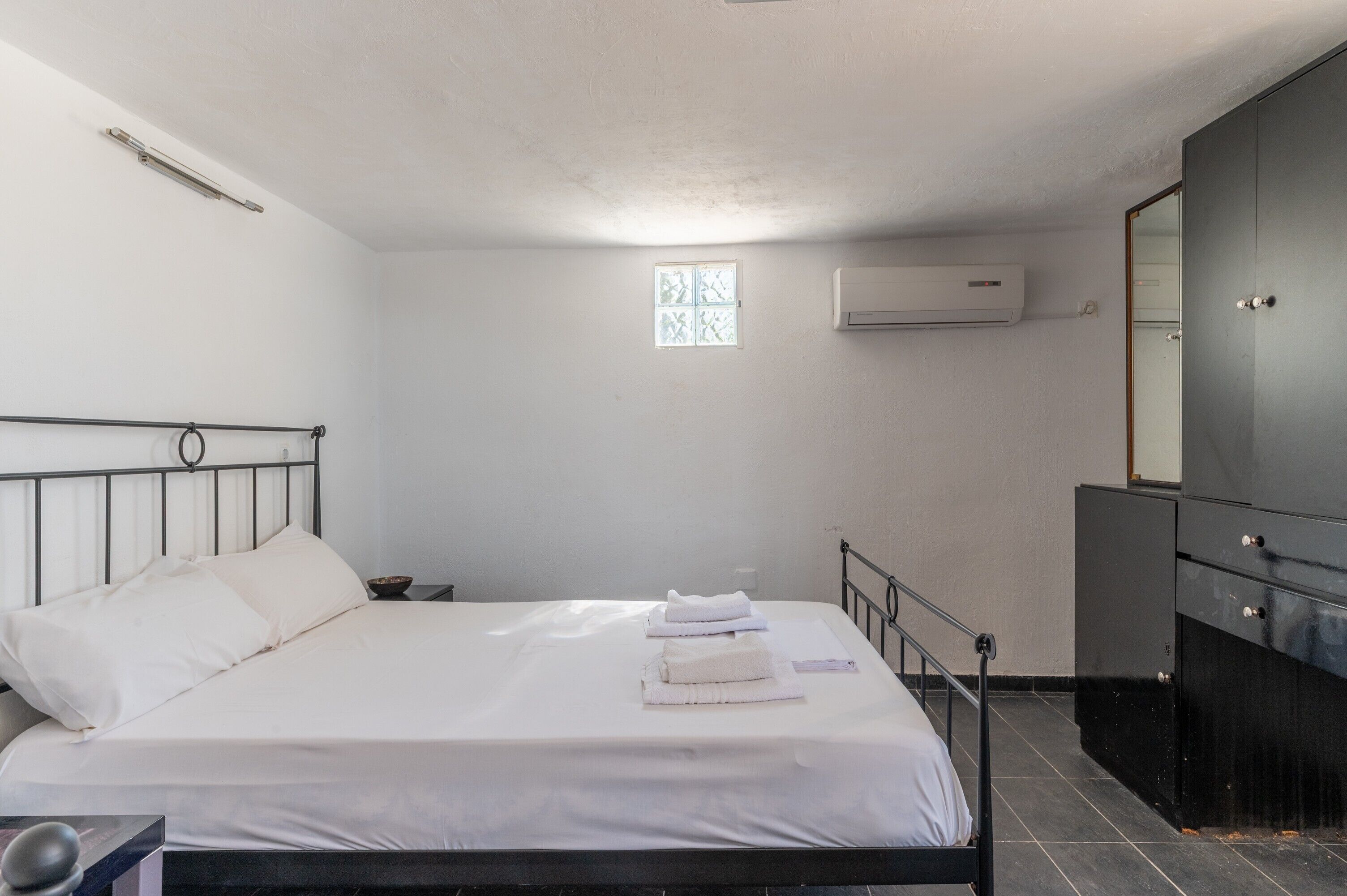 Een slaapkamer met een bed, een klein raam, een airconditioningapparaat, een spiegel en een donkere dressoir.