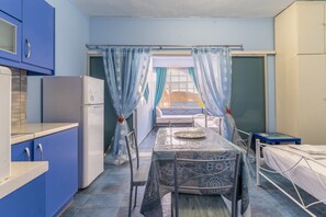 A compact kitchen with blue cabinets, a white refrigerator, a dining table with a patterned tablecloth, and a bed with a white mattress.