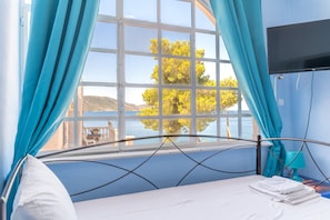 A room with a bed, a TV, a window with curtains, and a view of the sea and hills.
