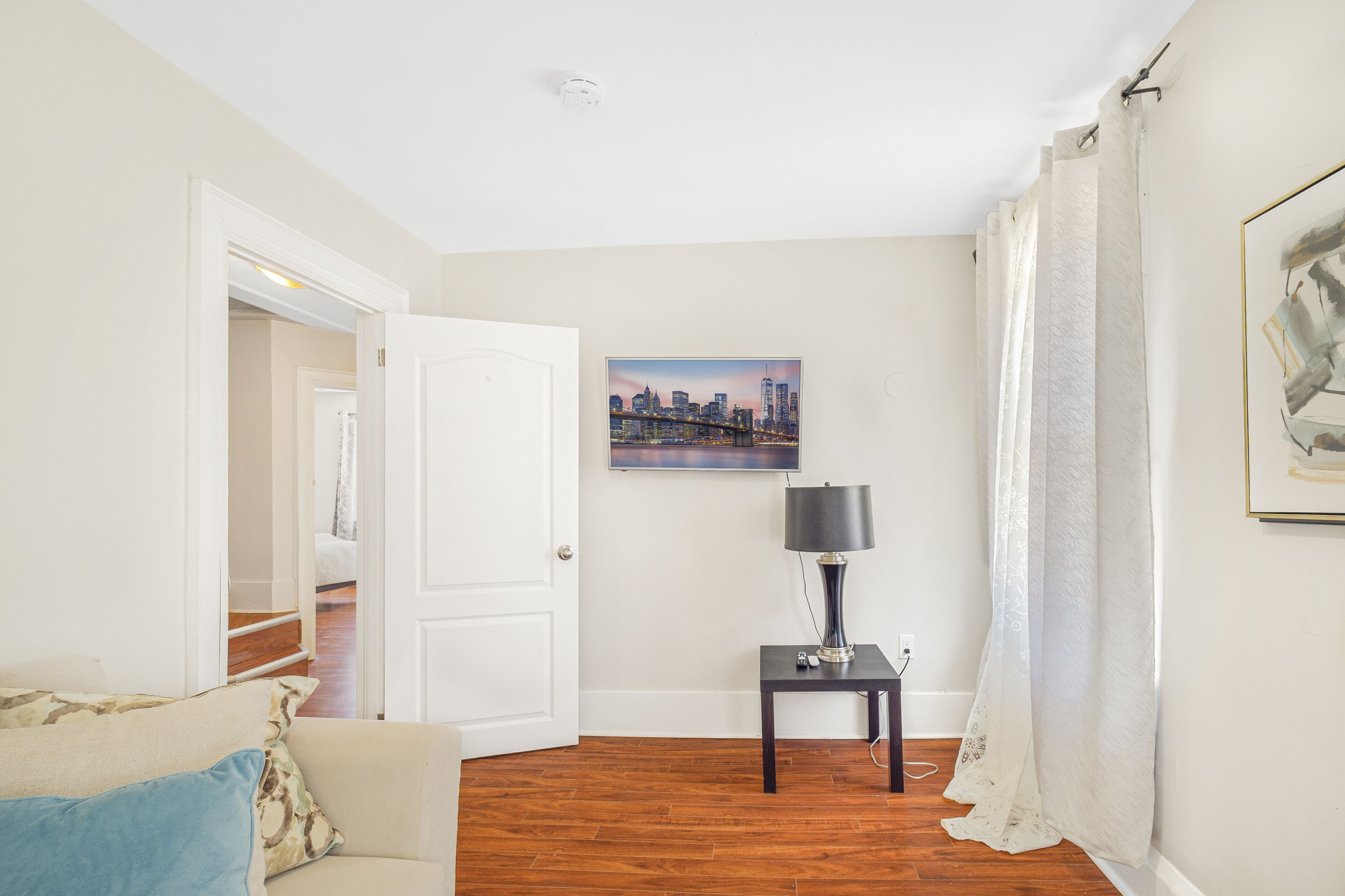 A living room with a wooden floor, a white door, a framed cityscape picture, a small table with a lamp, and a couch with cushions.