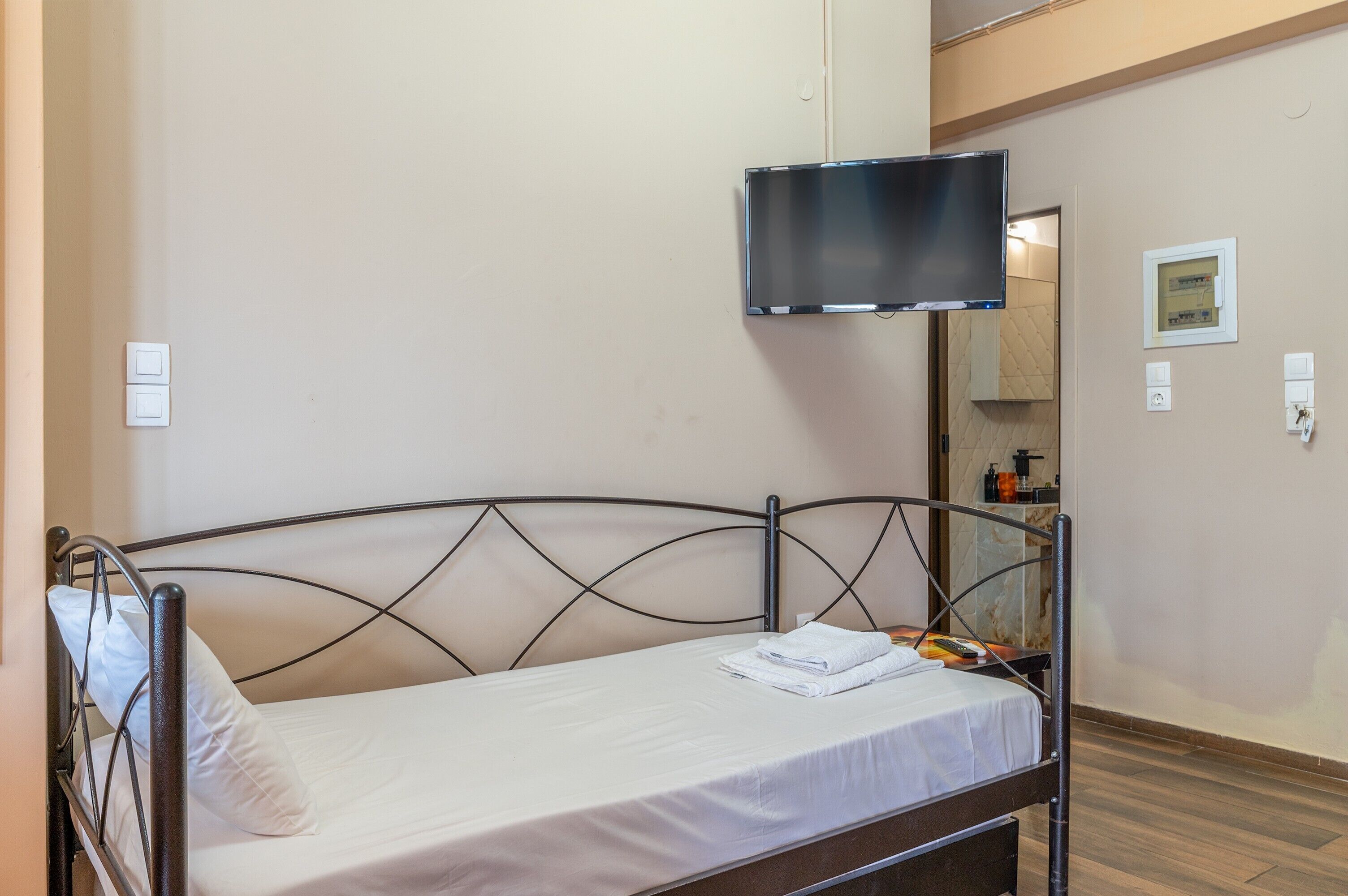 Ein Einzelbett mit Metallgestell, ein Flachbildfernseher an der Wand, ein kleiner Tisch mit Pflegeprodukten und eine Badezimmertür im Hintergrund.