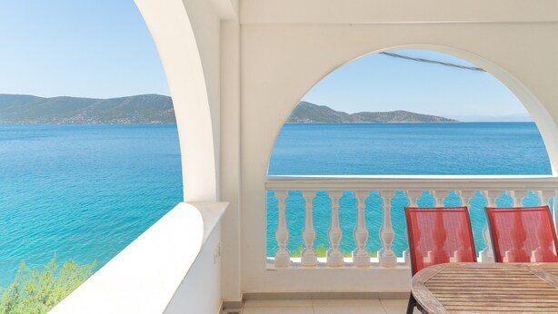 Ein Balkon mit Blick auf das Meer und die Berge, ein weißes Geländer und zwei rote Stühle.