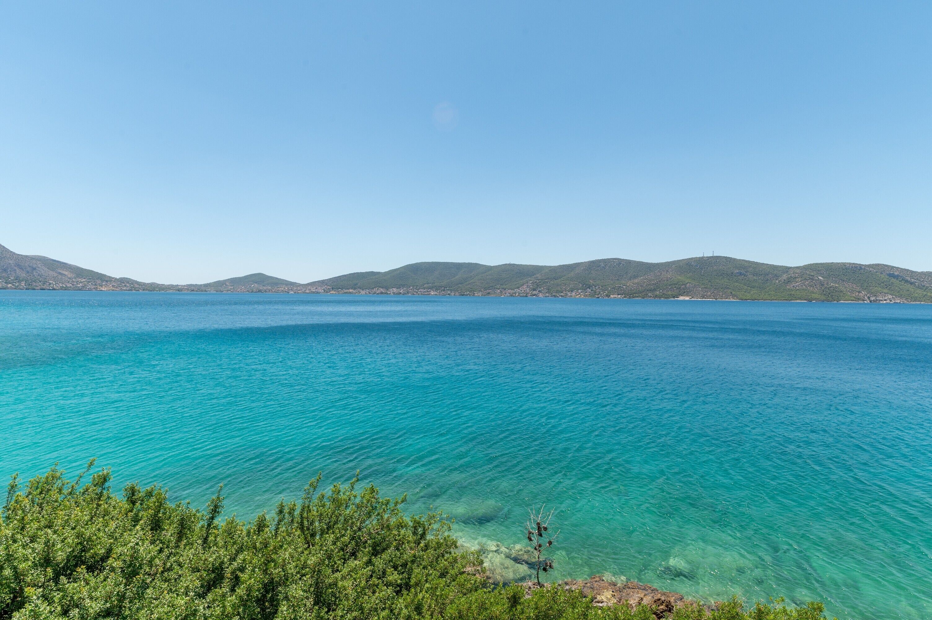 Paisagem costeira com águas turquesa cristalinas, colinas verdes ao fundo e vegetação densa em primeiro plano.