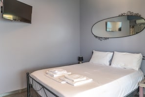 Un lit bien fait, avec du linge de lit et des serviettes blanches, un téléviseur fixé au mur, un miroir décoratif et une lampe de chevet.