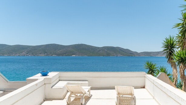 Eine Dachterrasse mit zwei weißen Liegestühlen und einer blauen Schale, mit Blick auf das klare, blaue Meer und sanfte Hügel in der Ferne.
