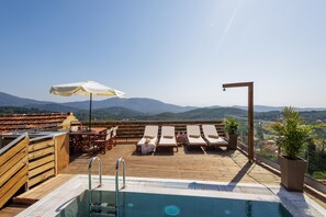 A rooftop pool with lounge chairs and a table, surrounded by wooden railings and a view of distant mountains.
