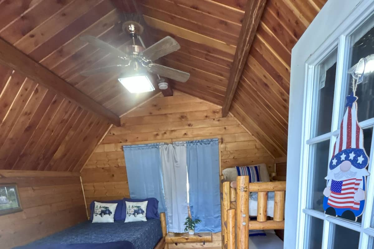 A cozy attic bedroom with a wooden ceiling, a single bed, a small table, and a window with a decorative gnome.