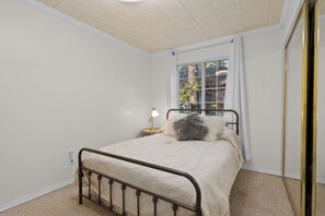 Une chambre à coucher avec un lit, une table de chevet, une lampe et une fenêtre donnant sur des arbres et des immeubles.