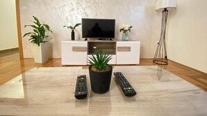 A modern living room with a flat-screen TV on a white cabinet, two remote controls, a potted plant, and a floor lamp.